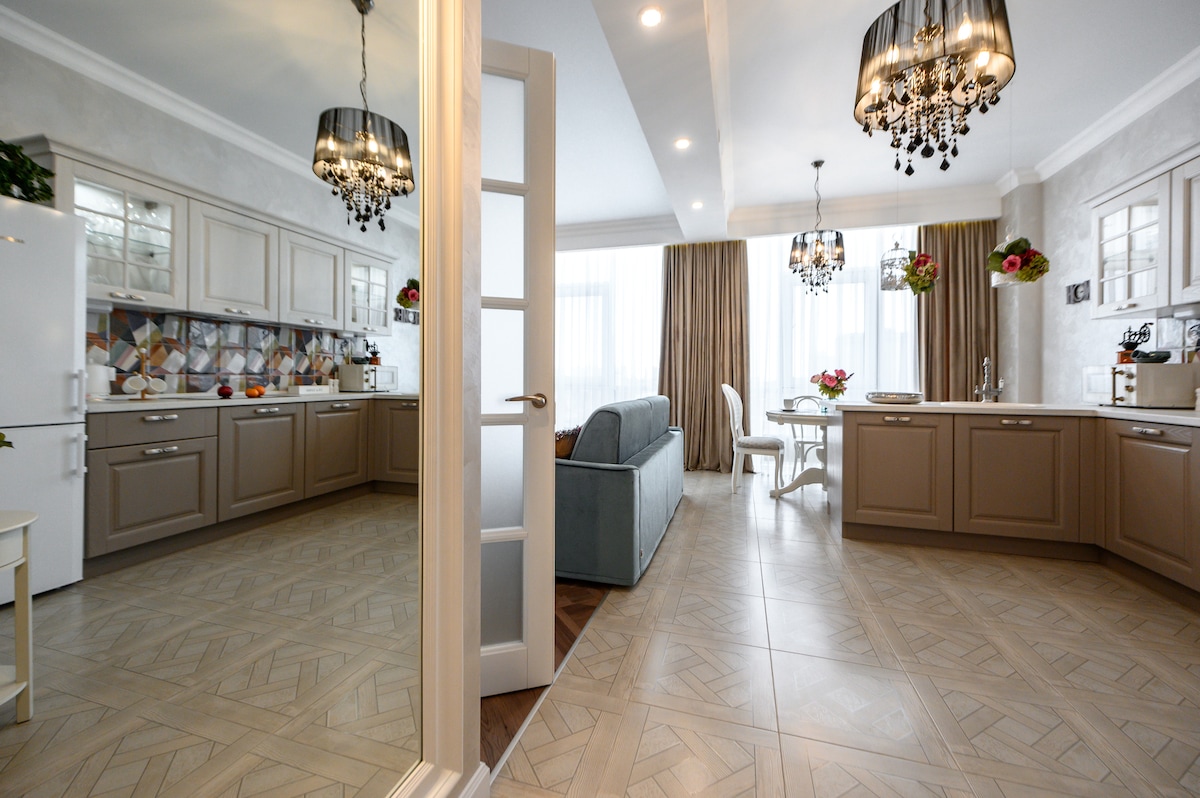 The open kitchen and living area are featured, showcasing warm neutral cabinetry and a dining table set for four. Natural light floods the space through large windows adorned with soft curtains, while decorative chandeliers hang from the ceiling, adding elegance to the room.