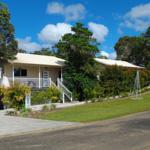 A light-colored cottage is surrounded by lush greenery, with a well-maintained lawn in the foreground. The entrance features a wide staircase leading to a welcoming verandah adorned with tropical plants, set against a backdrop of partly cloudy blue skies.