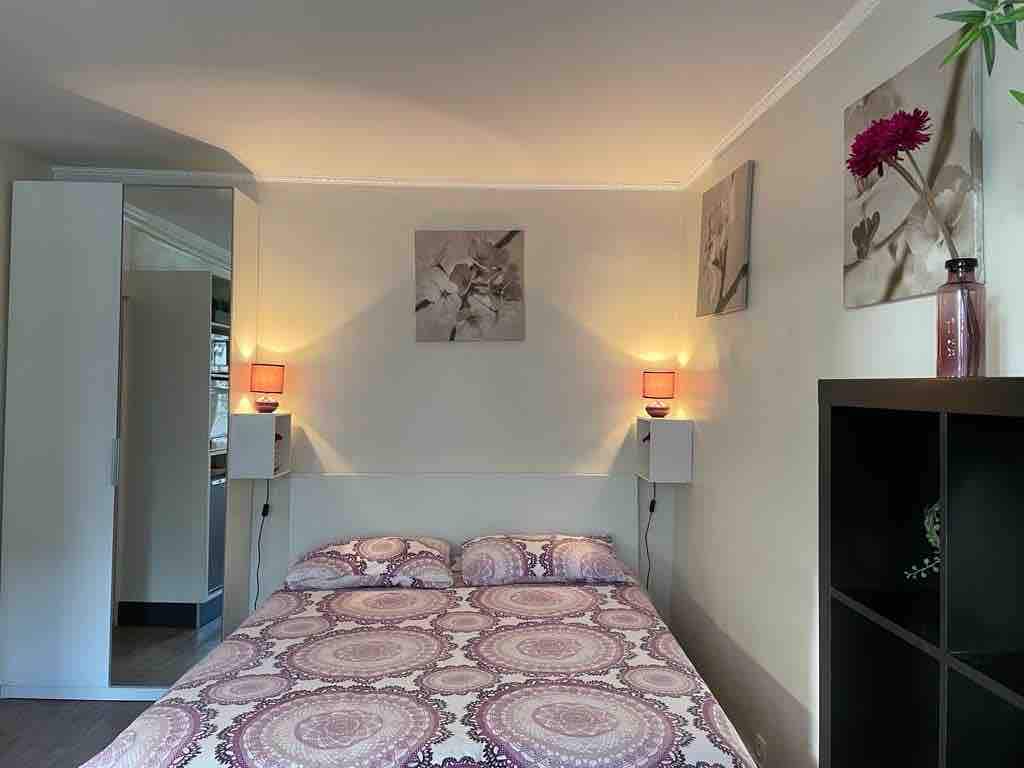 A spacious bedroom is presented, featuring a patterned bedspread and two bedside lamps providing a warm glow. Above the bed, decorative artwork is displayed on light-colored walls, and a wardrobe is visible to the left, offering storage solutions.