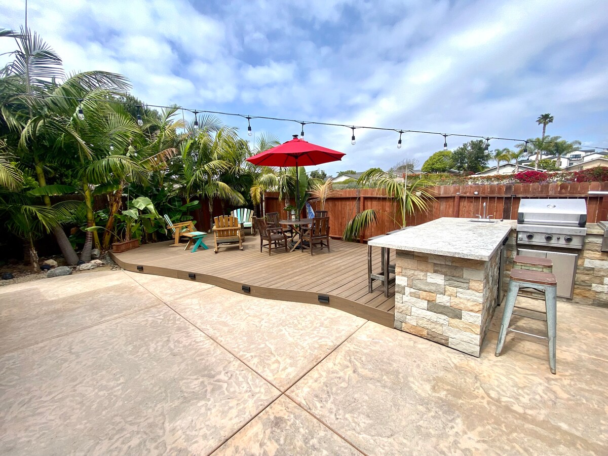 An outdoor living space is presented with a wooden deck surrounded by palm trees. A dining table with four chairs and a large red umbrella offers shade. An outdoor kitchen with a grill and counter space is included, enhancing the area for meal preparation and socializing.