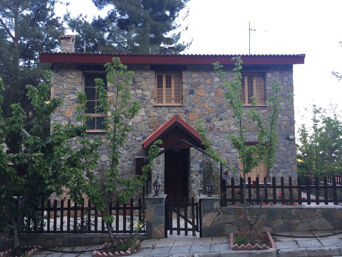 A two-story stone house is featured, showcasing wooden shutters and a welcoming front entrance. The structure is surrounded by greenery and a low stone fence, enhancing the outdoor experience. A paved pathway leads to the entrance, creating a charming and inviting approach.