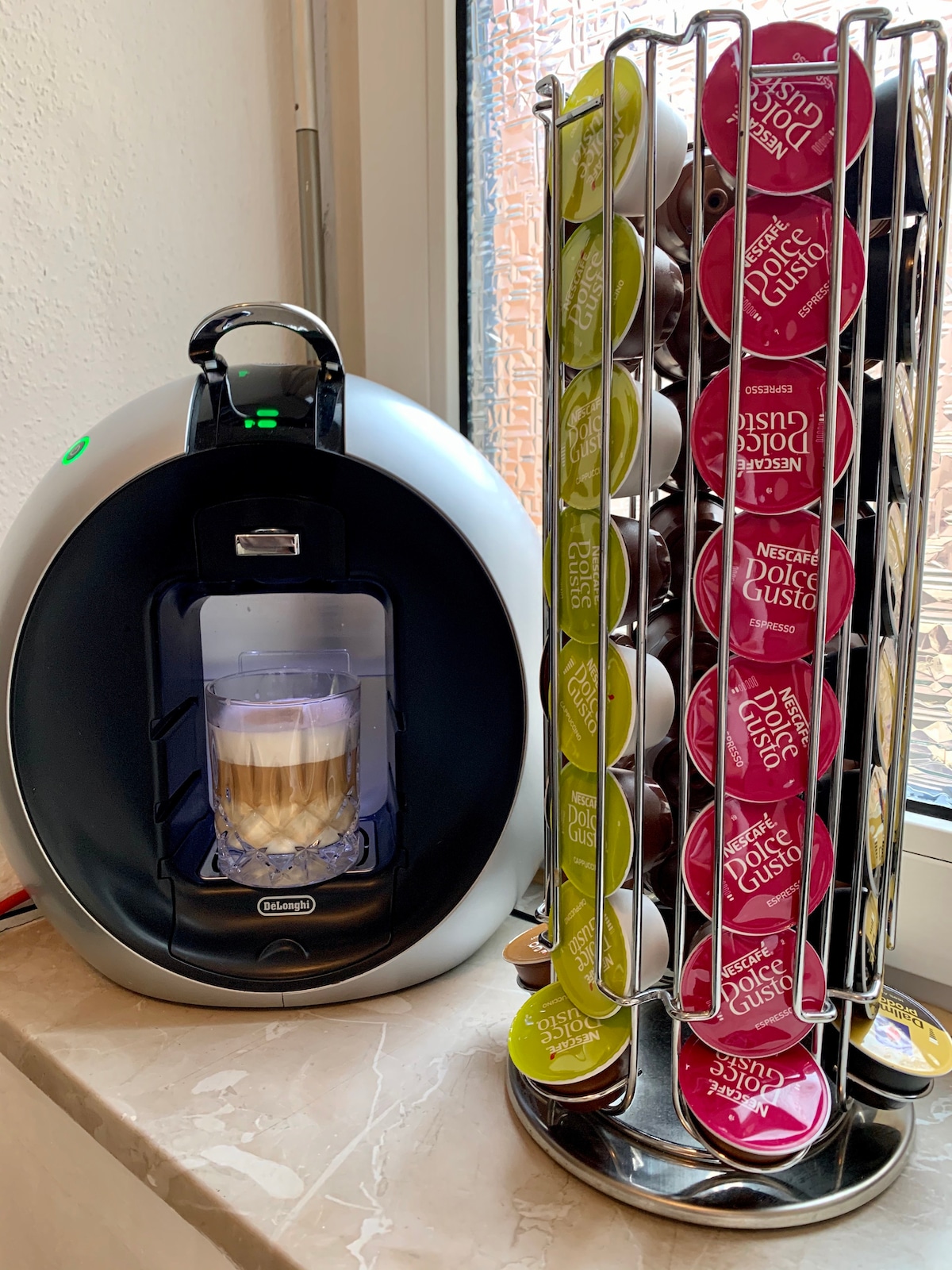 A coffee machine is positioned next to a rotating carousel of colorful coffee capsules. The machine is designed to brew various coffee styles and features a cup already filled with coffee. Natural light is visible, highlighting the countertop.