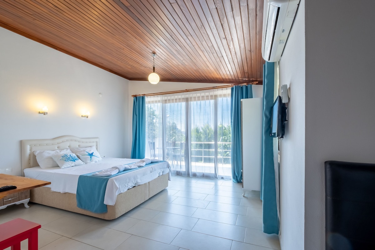 A bright and airy room features large windows with sheer curtains allowing natural light. A spacious bed is dressed in white linens with accent pillows. A wooden ceiling adds warmth, while a small red table and a black chair are visible against the tiled floor.