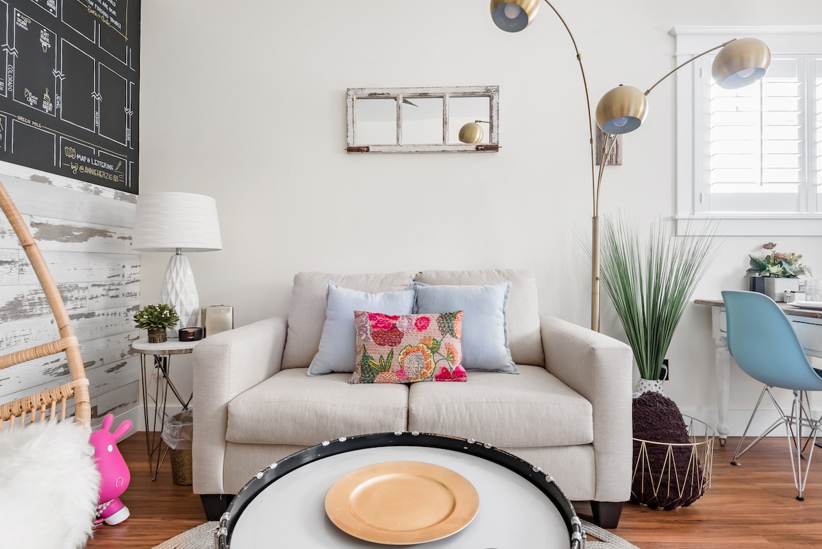 A cozy living area features a light-colored sofa adorned with decorative pillows. A modern floor lamp stands beside the sofa, and a round coffee table is set at the center. Vibrant greenery and wall decor add character to the welcoming space.