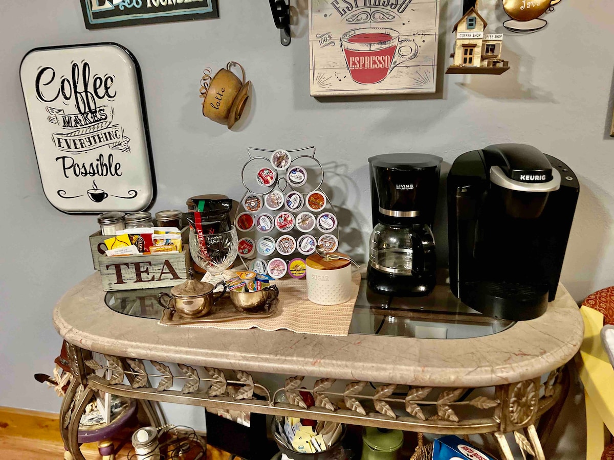 A charming coffee station is presented, featuring a modern Keurig machine and a traditional coffee maker. Decorative canisters labeled 'Tea' and 'Coffee' sit on a shelf above a colorful carousel of coffee pods. A sign reading 'Coffee makes everything possible' adds a welcoming touch.