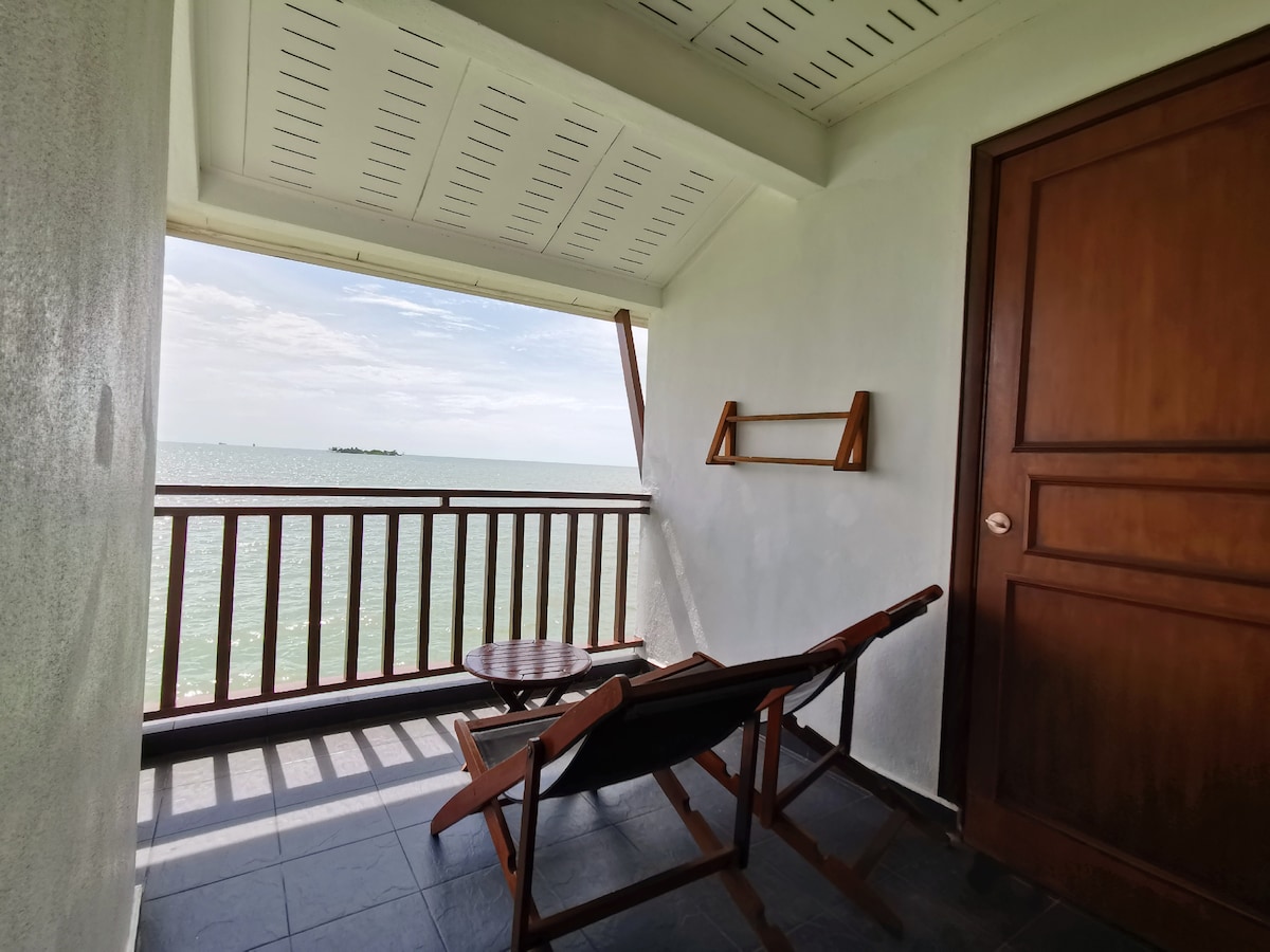 A private balcony is visible, featuring a wooden chair and a small table, both angled to overlook the sea. The serene water views are framed by an angled roof and beige walls, creating a peaceful retreat. A wooden door leads into the room.