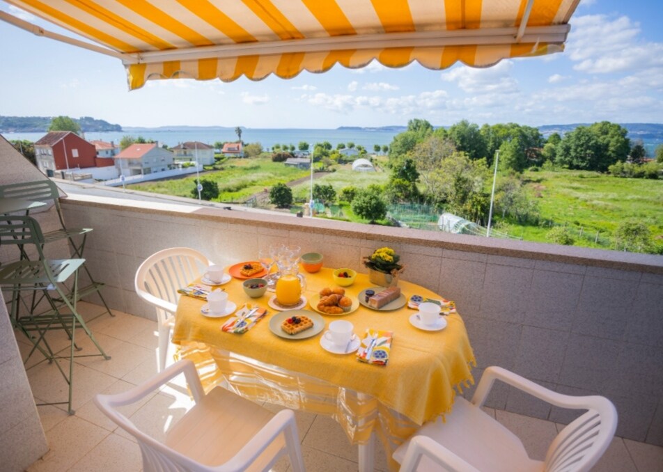 A balcony table is set for breakfast, featuring a yellow tablecloth and a variety of dishes, including pastries and drinks. The view includes distant water and greenery, framed by bright blue skies and scattered clouds. Light-colored chairs are arranged around the table.