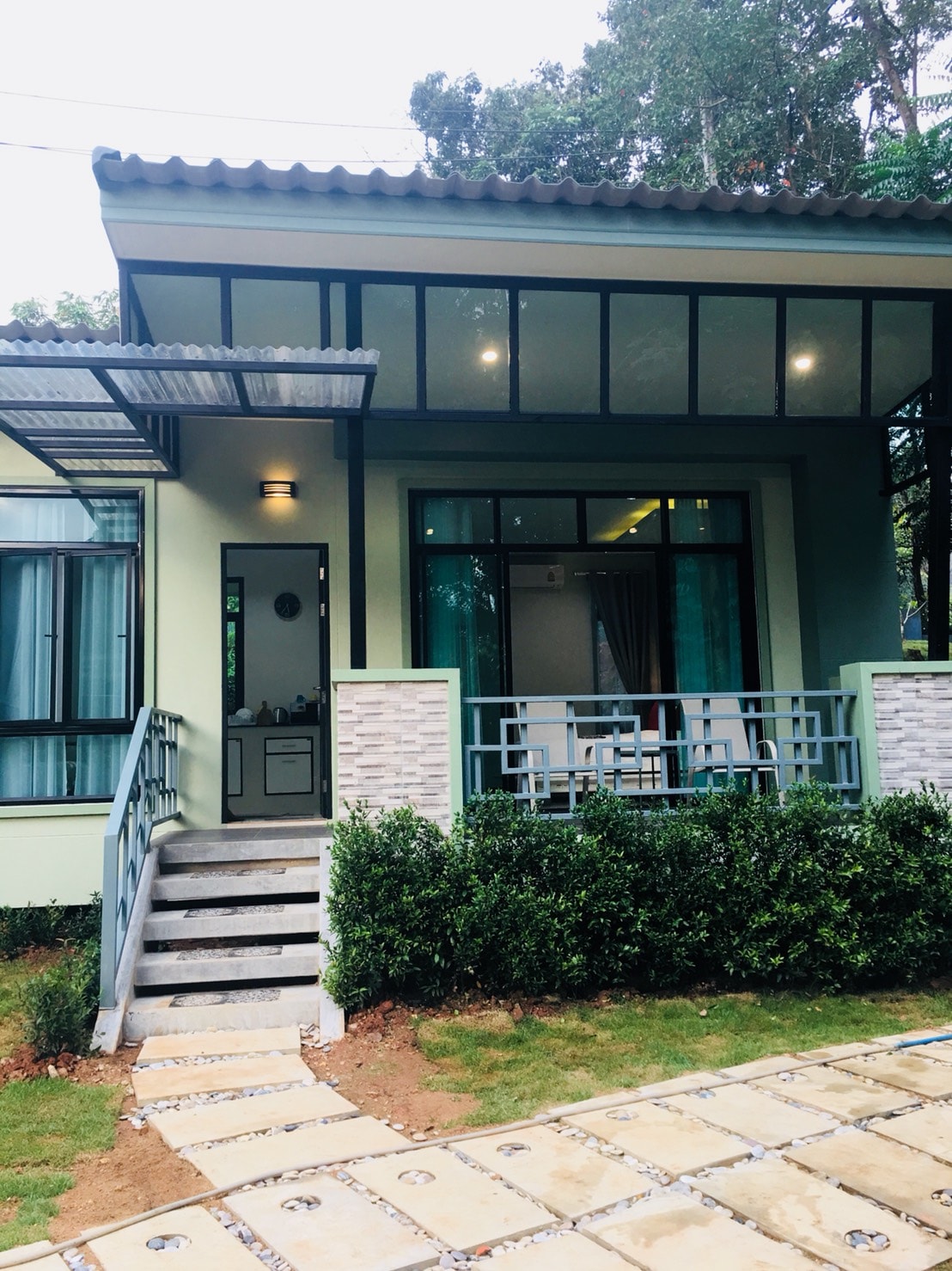 A contemporary house exterior is framed by large windows, allowing natural light to fill the interior. A small kitchenette is visible through the glass. A stone pathway leads to the entrance, bordered by well-maintained greenery, contributing to a welcoming atmosphere.