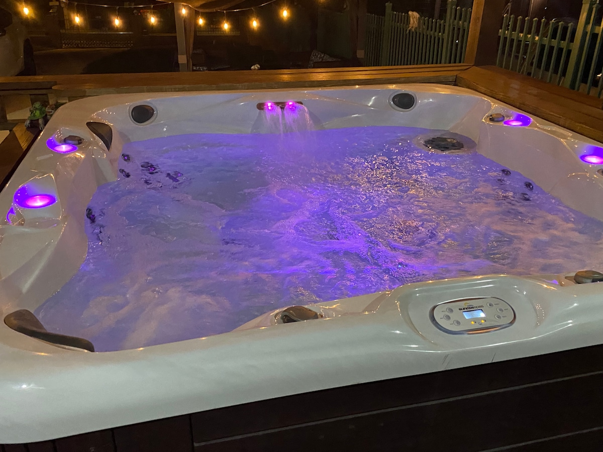 A spacious jacuzzi is illuminated with soft, colorful lights, creating a tranquil atmosphere. Bubbles gently rise in the warm water, surrounded by a wooden deck. The setting is enhanced by string lights in the background, providing a cozy ambiance for relaxation.