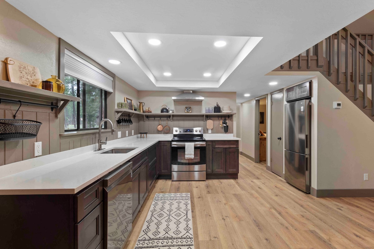 A modern kitchen features sleek dark cabinets and light countertops, complemented by stainless steel appliances. A geometric area rug lies on the warm wood floor, while natural light filters through a window above the sink, enhancing the functional layout.