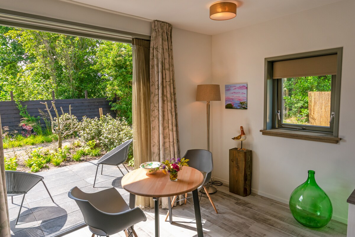 A bright and airy seating area features a round table with two chairs, complemented by a large green bottle and a lamp. Large sliding doors open to a landscaped garden filled with greenery, creating a peaceful outdoor view.