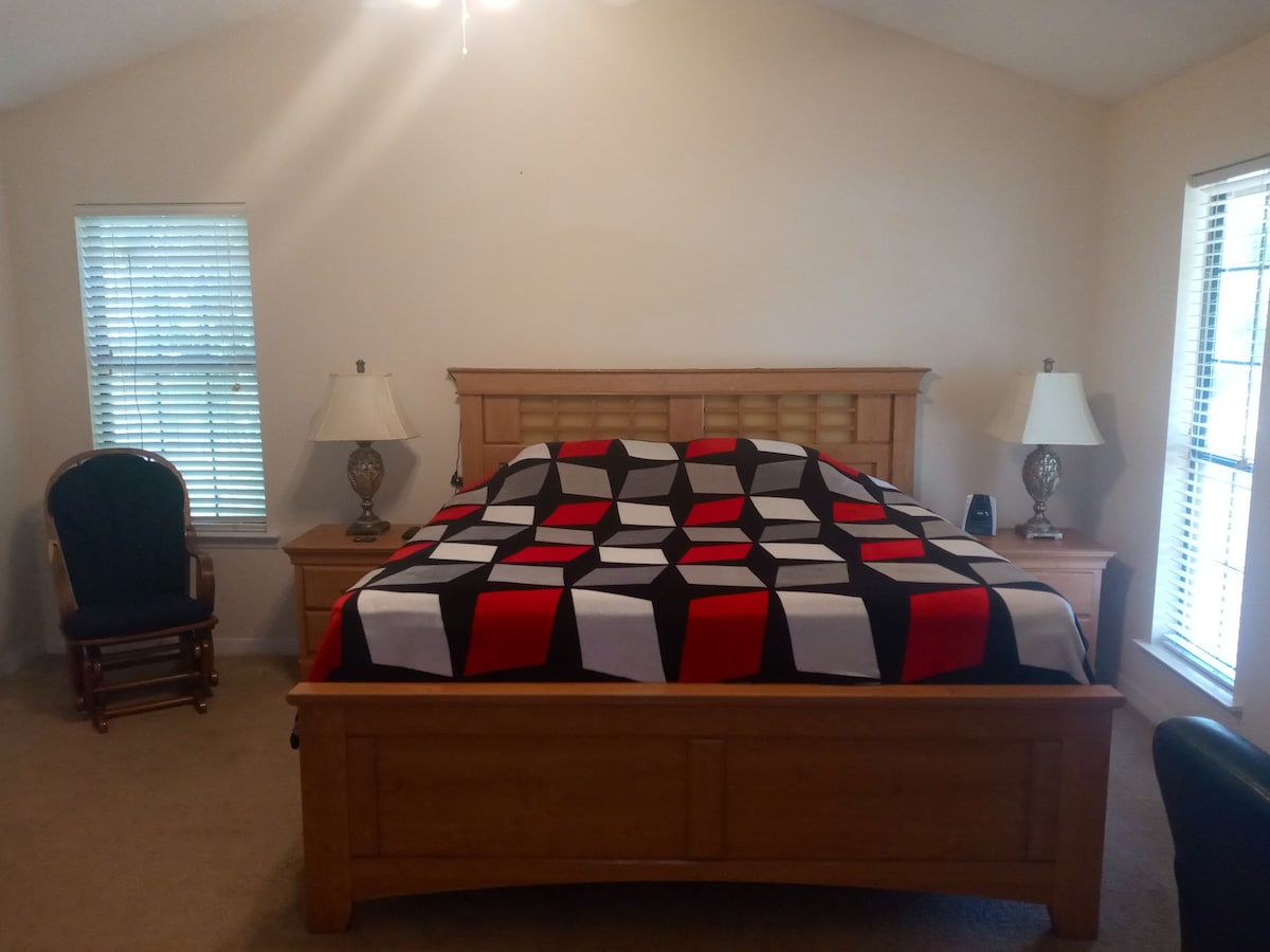 A spacious bedroom features a large wooden bed topped with a contrasting black, white, and red patterned blanket. Two bedside lamps provide soft lighting on either side, and windows with blinds allow natural light to brighten the room.