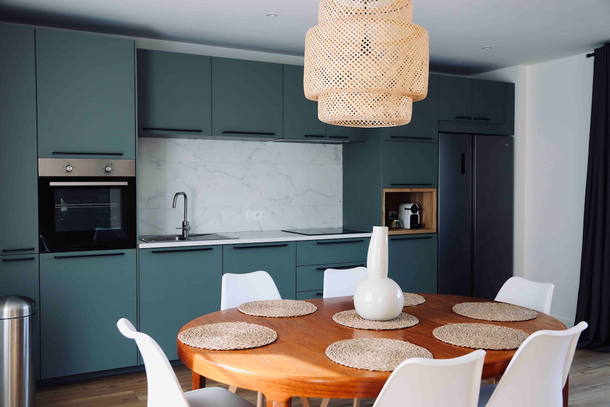 A modern kitchen is presented, featuring teal cabinetry and a marble backsplash. A round wooden table is set at the center, adorned with woven placemats and a minimalist vase. A pendant light provides soft illumination, and appliances are neatly integrated into the design.