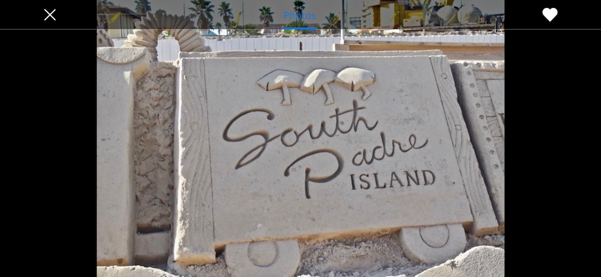 A sand sculpture displaying the words 'South Padre Island' is featured, accented with decorative elements such as seashells and palm trees in the background. This sign reflects the beach environment and serves as a welcoming marker for visitors to the island.