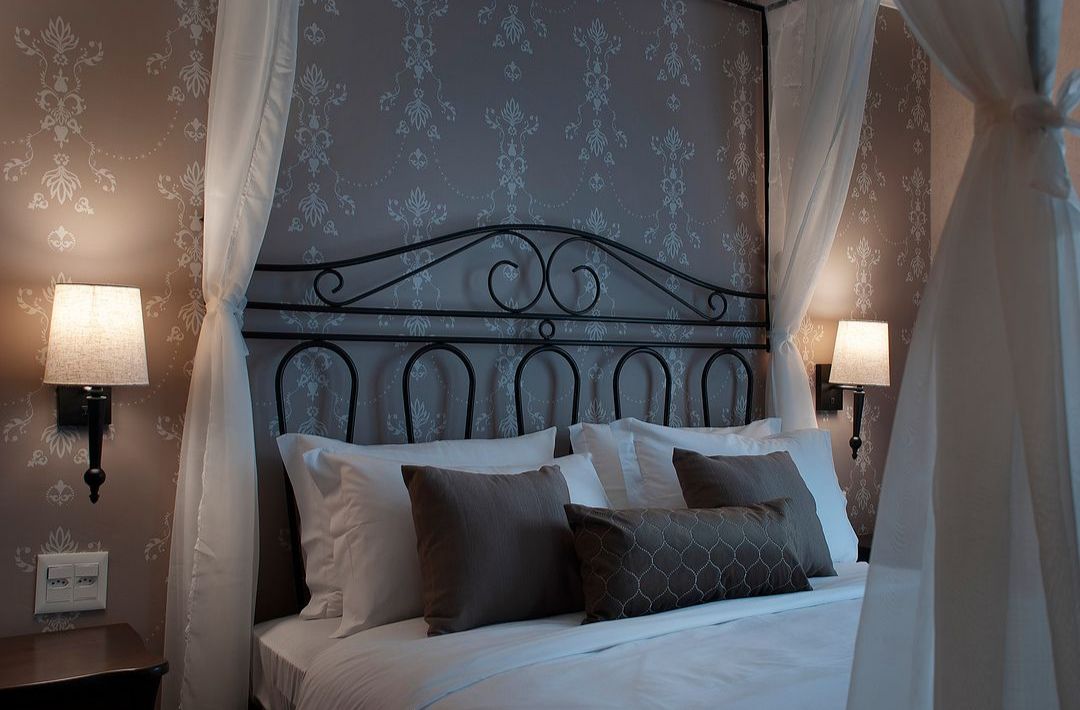 A cozy bedroom features a four-poster bed dressed in crisp white linens, with plush pillows in muted tones. Decorative wall sconces provide soft lighting, illuminating the intricately patterned wallpaper in shades of gray, creating a serene ambiance.