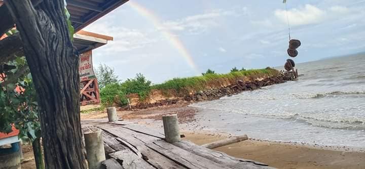 A wooden pathway leads to the sandy shore, where gentle waves meet the beach. Lush greenery lines the background, and a rainbow arches over the horizon, creating a serene coastal view.