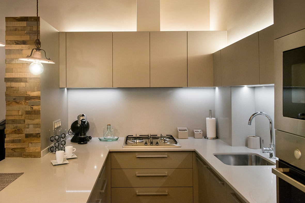 A modern kitchen is featured, showcasing sleek, light-colored cabinetry and a polished countertop. Cooking appliances, including a stove and oven, are visible. Task lighting emphasizes the space, while an eclectic wall adds texture. Kitchenware such as a coffee maker and utensils are neatly arranged.