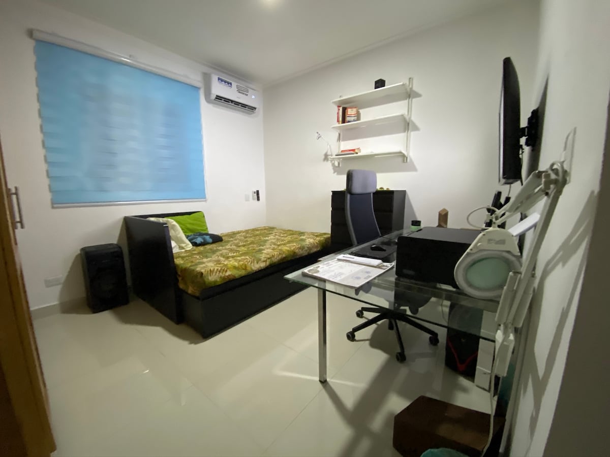 A compact room features a well-organized workspace with a glass desk and a black office chair. A spacious bed, adorned with a patterned green and brown blanket, sits against the wall. A shelf holds books and decorative items, while a window with light blue blinds allows natural light in.