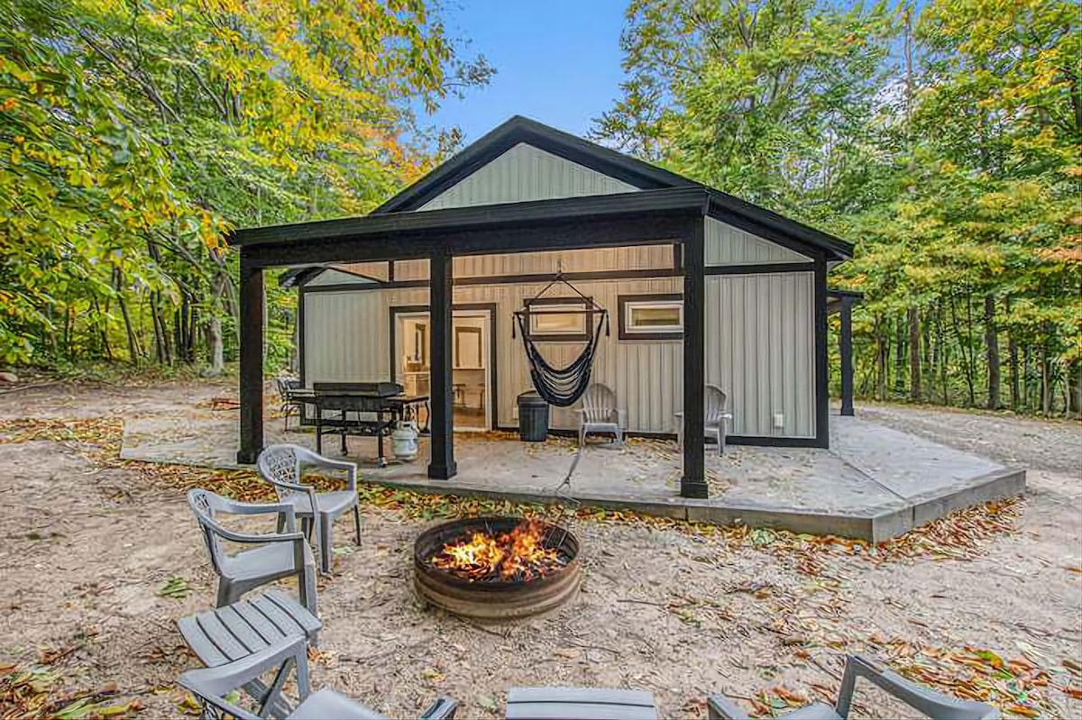 An outdoor gathering area features a fire pit surrounded by several gray chairs. A covered patio showcases a dining table and a hammock, with a backdrop of colorful autumn trees that provide privacy and a natural setting.