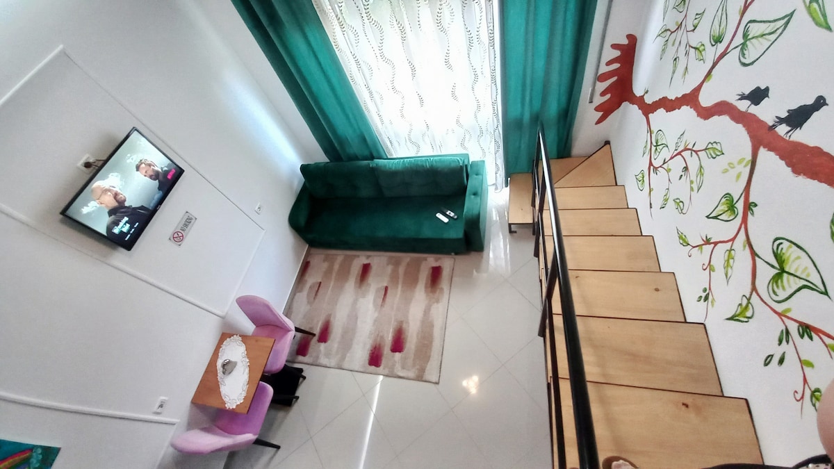 A well-lit living space is visible, featuring a green sofa adjacent to a window with sheer curtains. A simple dining table with two chairs is present, along with a decorative area rug. Stairs lead to an upper level with a tree mural adding a touch of creativity.