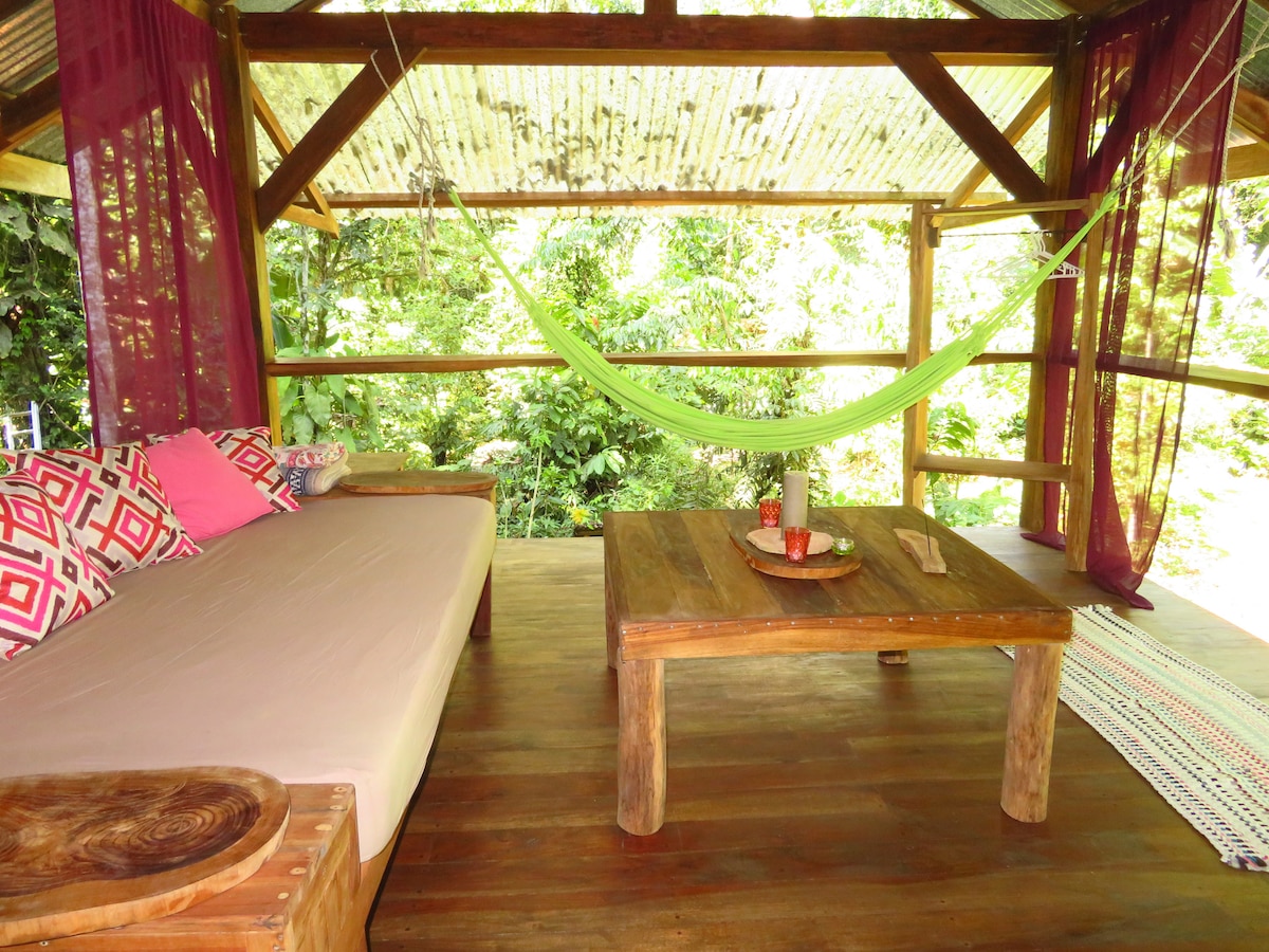 A shaded outdoor seating area is presented, featuring a spacious bed-like daybed with decorative pillows. A bright green hammock hangs above a rustic wooden table, which is adorned with simple decor items. Lush greenery surrounds the area, enhancing the natural ambiance.