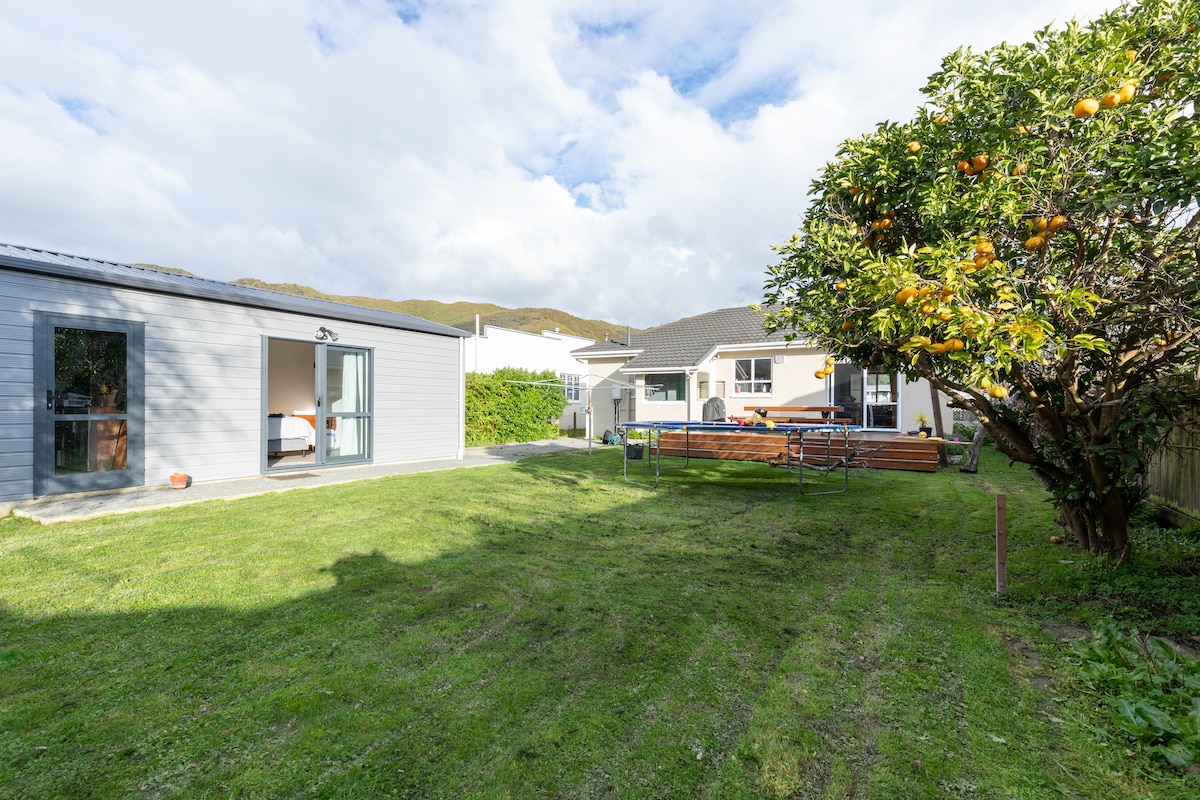 A spacious backyard is highlighted, featuring a well-maintained lawn with a grapefruit tree and a wooden table. In the background, the studio apartment is visible on the left, along with a main house and a deck area, inviting opportunities for relaxation.