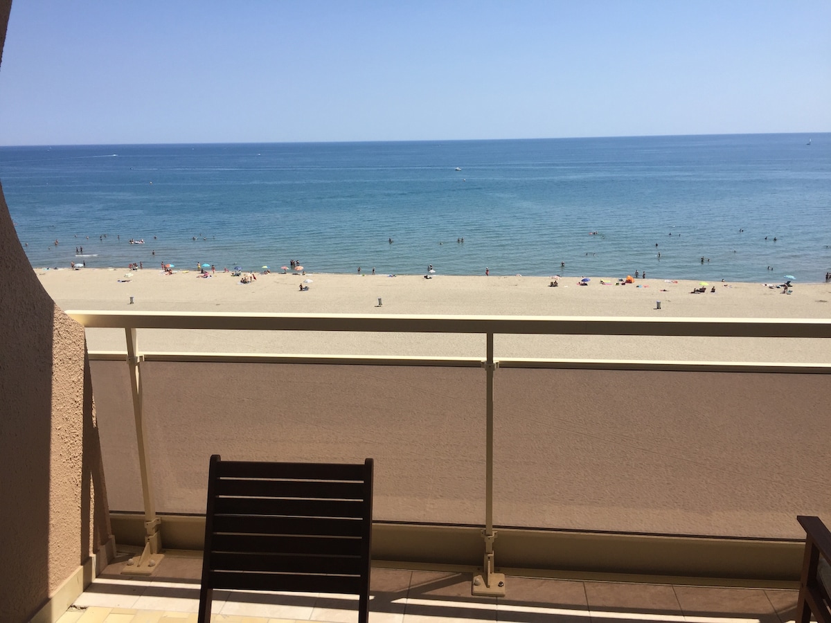 A spacious terrace showcases a stunning view of the sandy beach and calm sea. Sunbathers and beachgoers can be seen on the shoreline, while the clear blue sky enhances the coastal atmosphere. A wooden chair positioned on the terrace invites relaxation.
