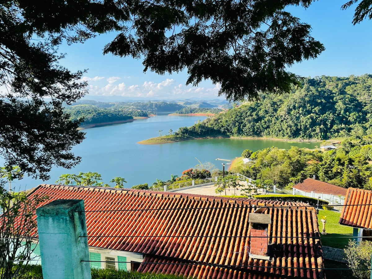 A panoramic view showcases a tranquil lake surrounded by lush greenery and distant hills. The foreground features a rustic red-tiled roof, emphasizing the connection between the landscape and the property. Bright blue skies and scattered clouds enhance the serene atmosphere of the scene.
