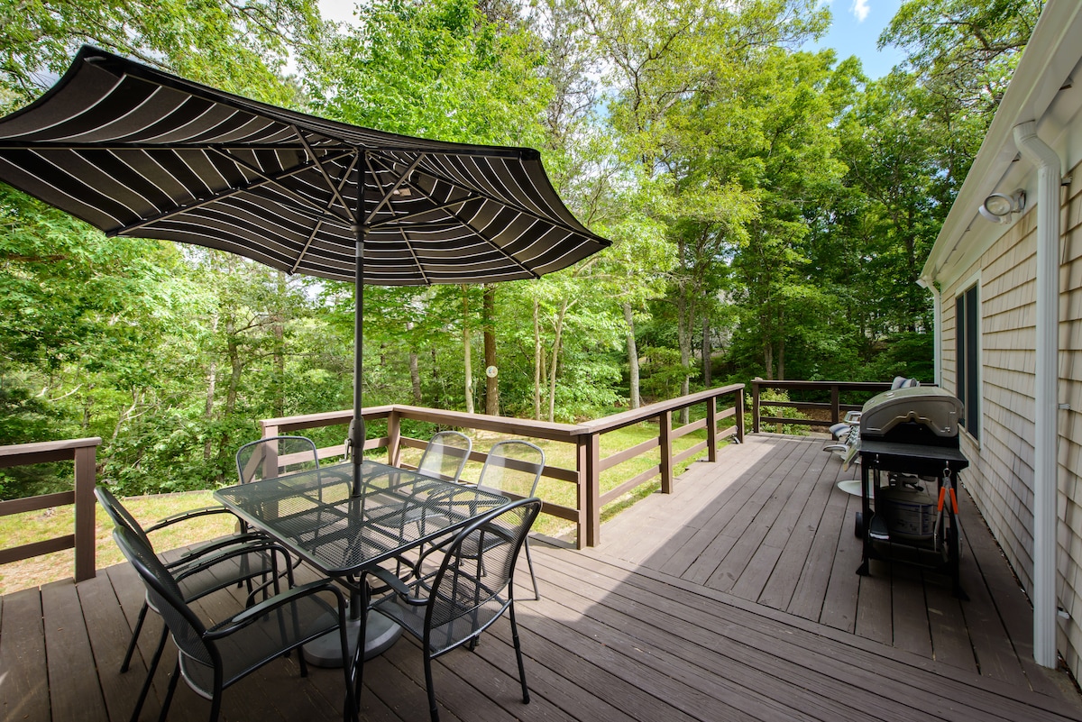 A spacious deck is surrounded by lush greenery, featuring a large table with six metal chairs beneath a striped umbrella. A gas grill is positioned nearby, creating an inviting outdoor space for dining and relaxation.