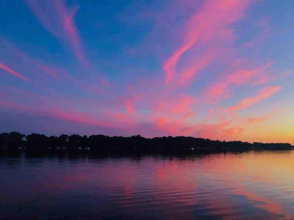 A tranquil lakeside view captures a sky painted with vibrant pink and purple hues at sunset. The calm water reflects the colors, creating a serene ambiance. A forested shoreline is visible in the distance, framing the picturesque scene.