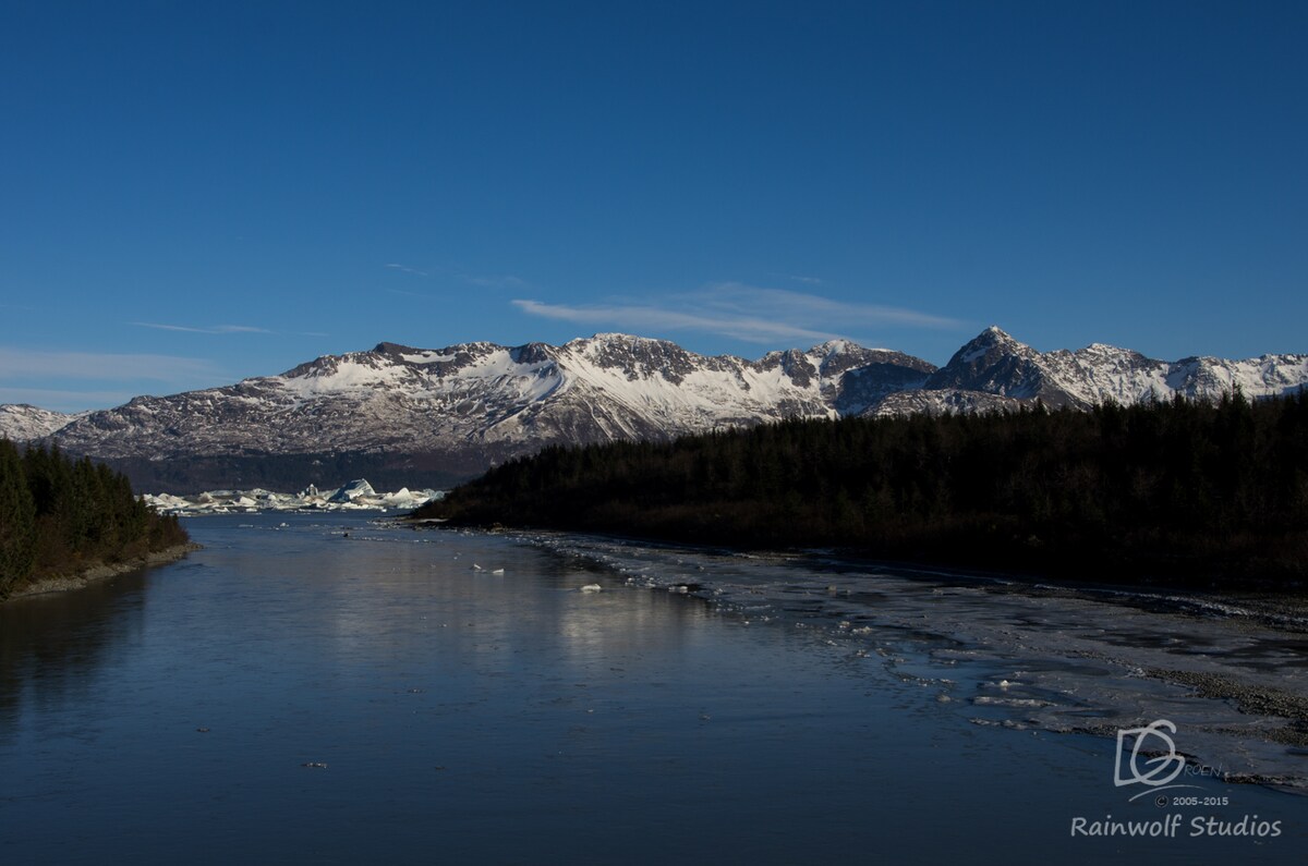 A serene landscape is displayed, featuring a calm river reflecting the bright blue sky. Snow-capped mountains rise majestically in the background, while patches of greenery line the riverbanks, creating a contrast against the icy blue hues of the water.