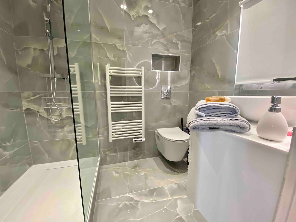 A modern bathroom features sleek grey marble walls and a glass shower stall. A white towel radiator stands beside the shower, while neatly stacked towels are arranged on the countertop. Natural light filters in through a frosted window, providing a bright and fresh ambiance.