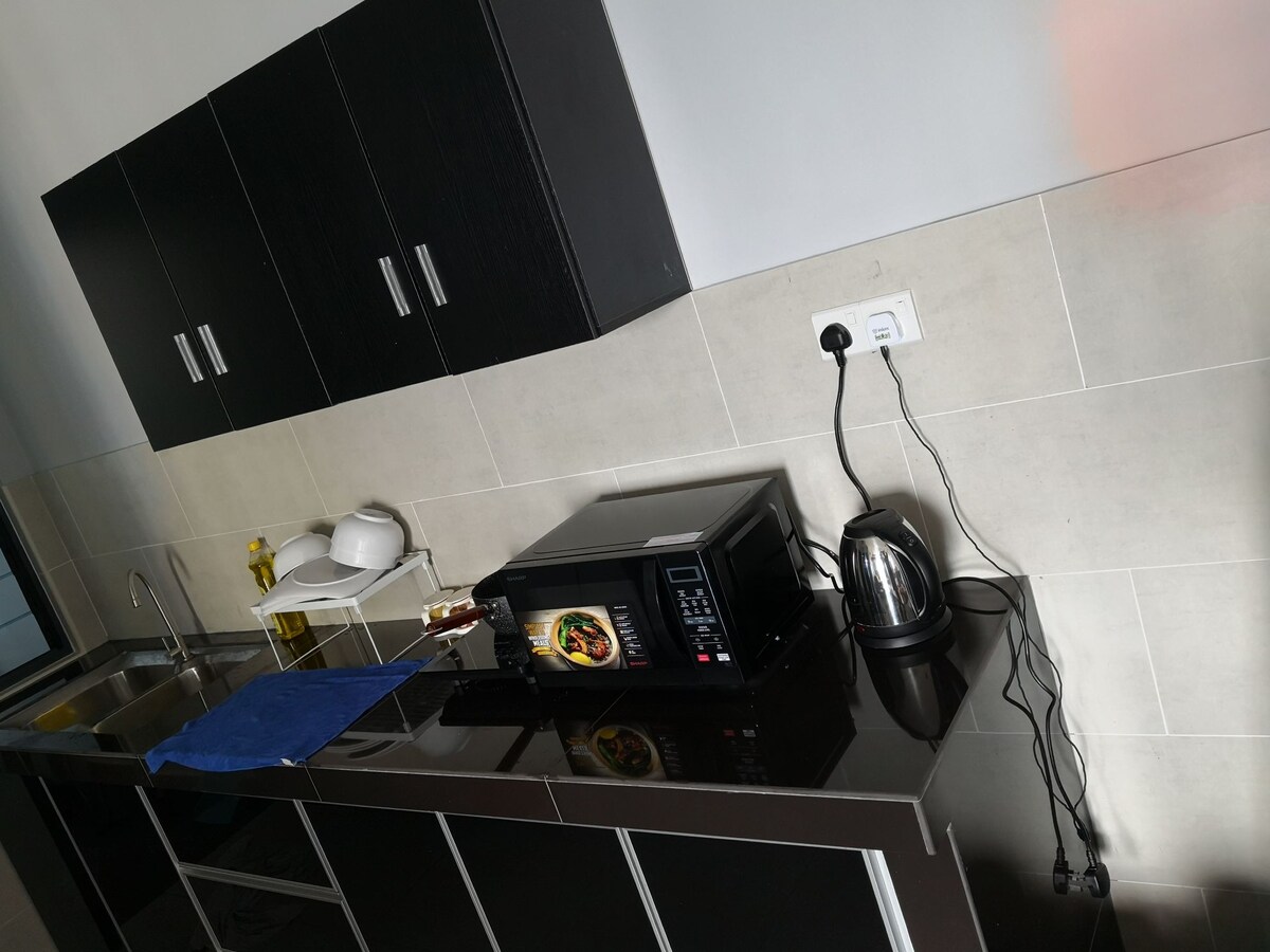 A modern kitchen with sleek black cabinetry and a smooth countertop is shown. A microwave and electric kettle are present, along with essential cooking utensils. Natural light brightens the space, highlighting the neutral-toned backsplash and stainless steel sink.