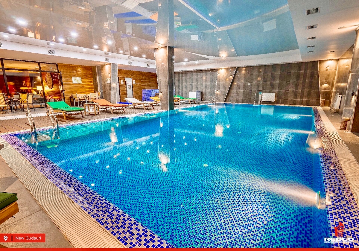 An indoor pool features a mosaic tile design with vibrant blue hues. Lounge chairs are arranged along the pool's edge, and soft lighting enhances the tranquil atmosphere. The spacious area is complemented by large windows allowing for natural light.
