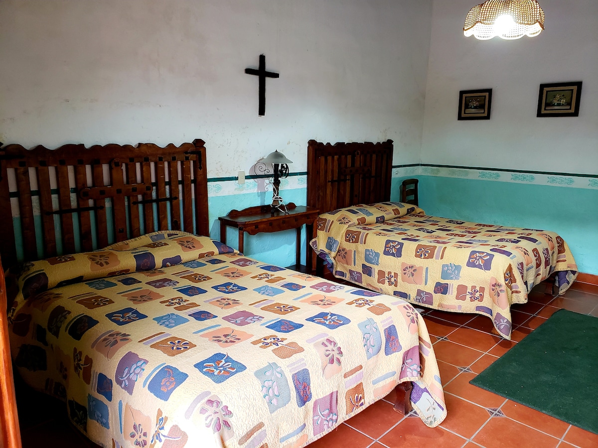 The room features two spacious beds adorned with colorful patterned blankets. A wooden lamp is placed on a small table beside one bed, while a simple chair and framed pictures are visible on the walls, adding a personal touch to the space.