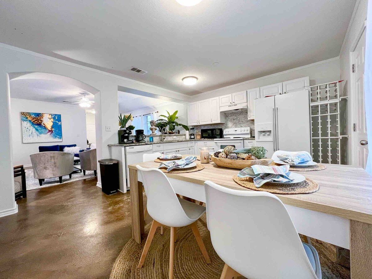 An open dining area is shown with a wooden table set for four, featuring decorative items and placemats. The adjoining kitchen is equipped with modern appliances, including a stove and refrigerator. Bright lighting illuminates the inviting space, seamlessly connecting to the living area beyond.