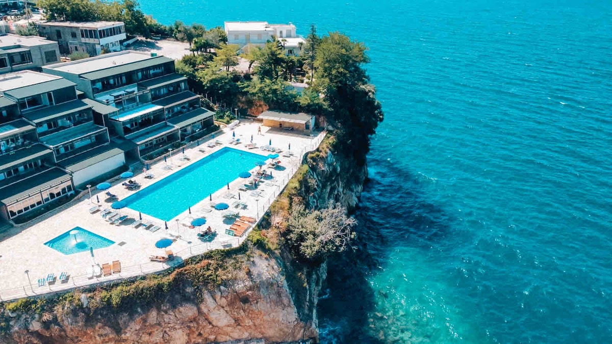 A seawater pool is surrounded by sun loungers and umbrellas, positioned on a cliff edge overlooking a vibrant blue sea. Green vegetation contrasts with the natural rock formations, and several buildings are visible in the background, enhancing the coastal setting.
