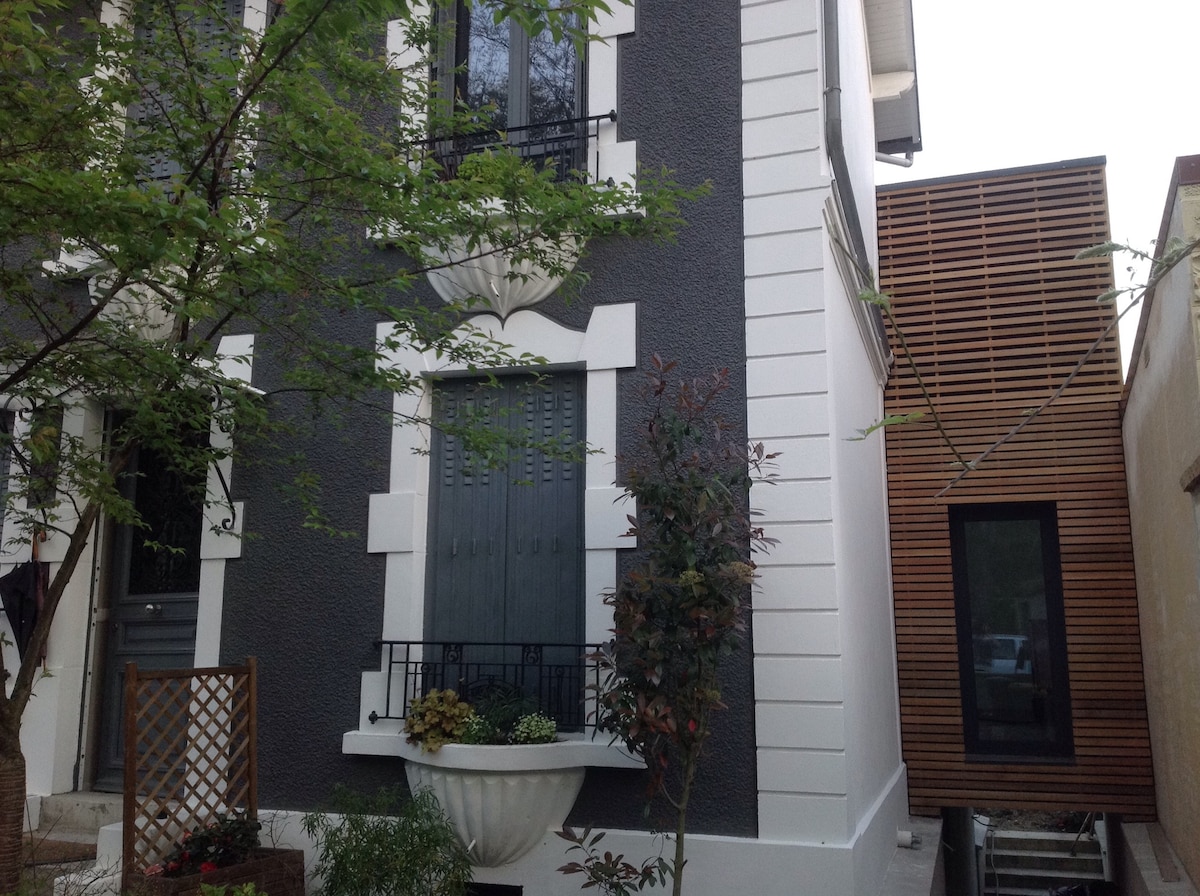The image depicts a two-tone building with distinctive architectural details. A gray facade features white trimming around the windows and a decorative planter. Adjacent to it, a modern structure with wooden slats and large windows complements the classic design, surrounded by greenery.