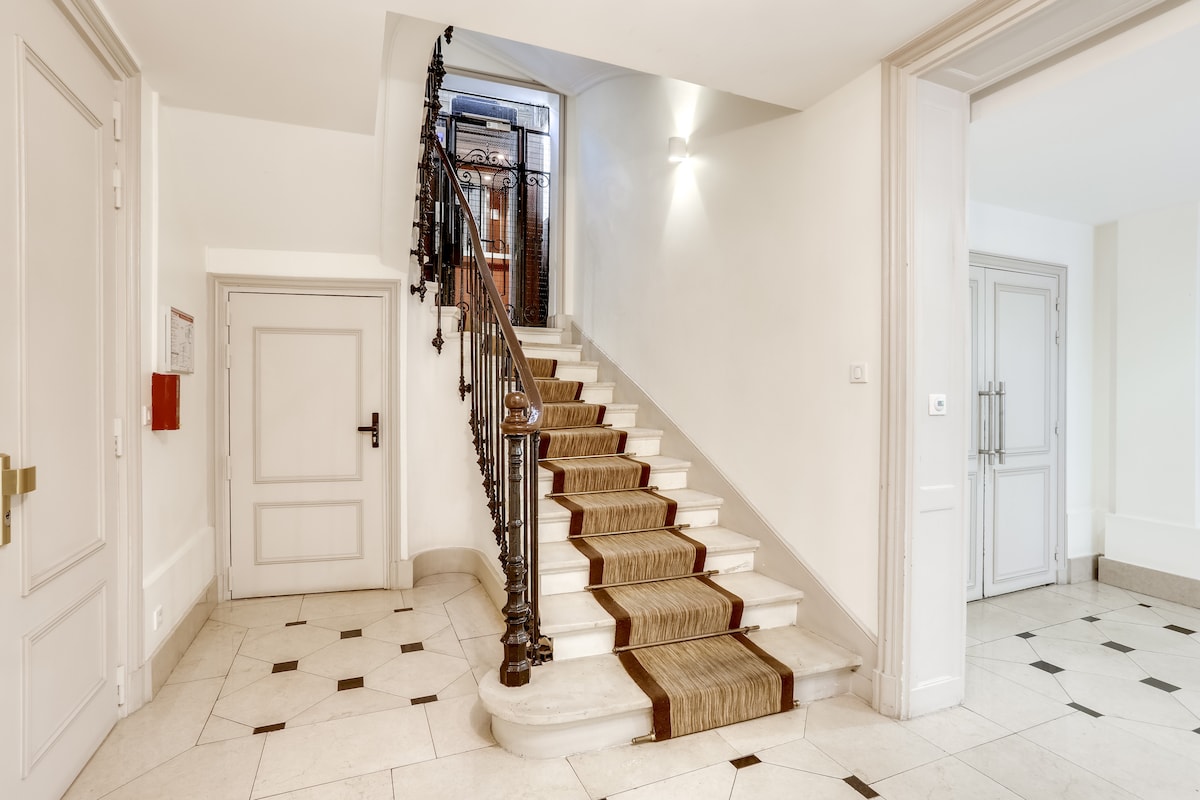 An elegant staircase is lined with a well-maintained runner, leading from a bright entryway to the upper levels. The walls are finished with light tones, and decorative moldings frame the spacious area. A subtle lighting installation enhances the ambiance.