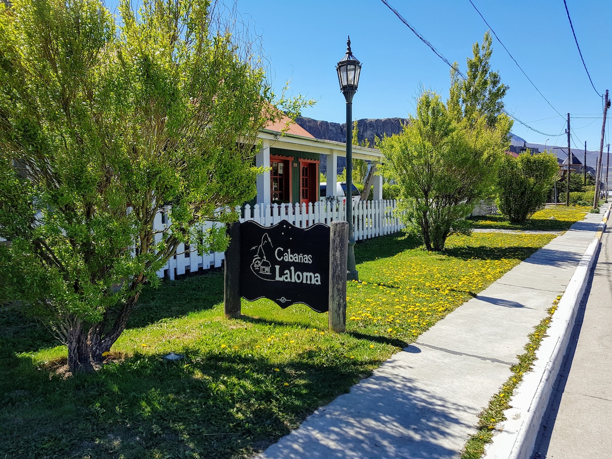 A welcoming entrance to Cabañas La Loma is captured, featuring a sturdy wooden sign surrounded by lush green grass and blooming dandelions. Neatly trimmed bushes frame the space, complemented by a charming lamppost and a white picket fence marking the boundary.