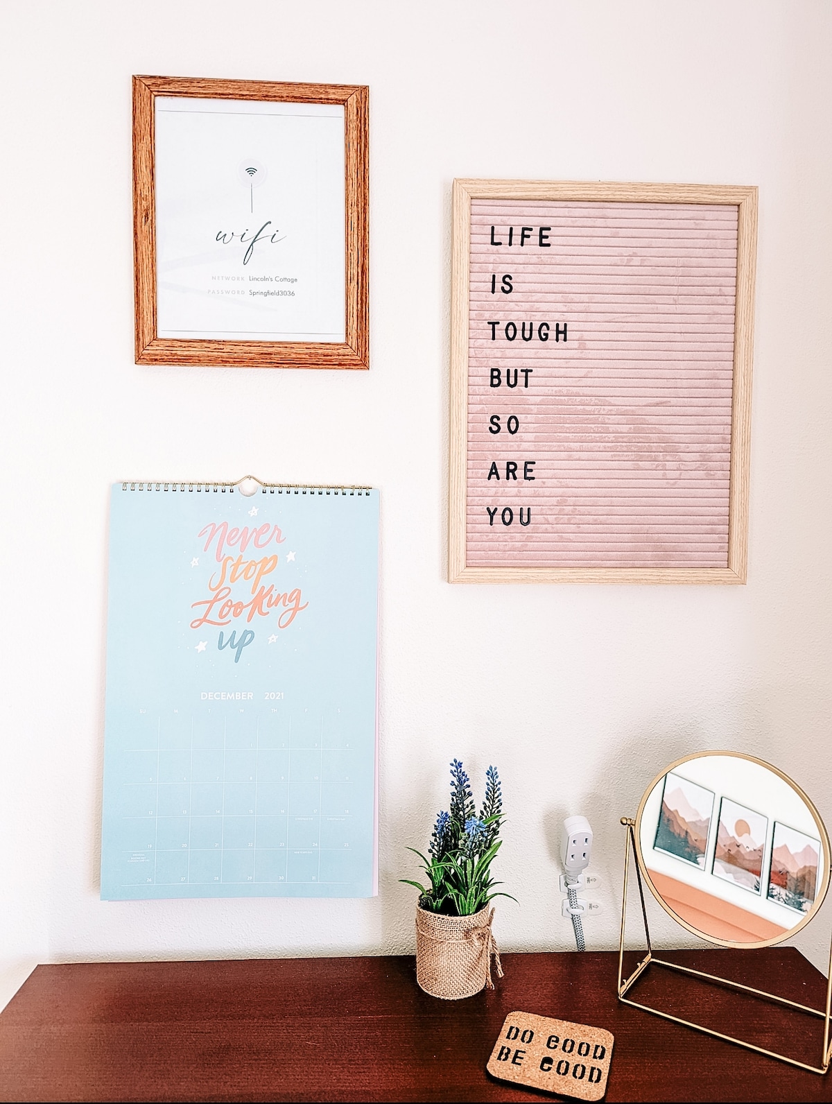A wall display featuring three framed items, including a print labeled 'wife', a letter board with motivational text, and a calendar marked with 'Never Stop Looking Up.' A small potted plant is positioned beside a round mirror on a wooden surface.