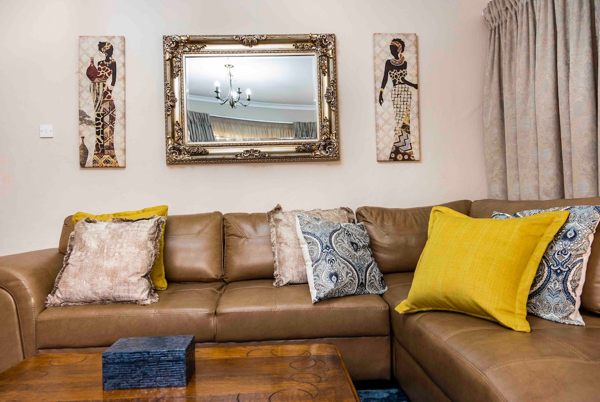 A leather sectional sofa is arranged around a wooden coffee table, adorned with decorative cushions in various patterns and colors. A vintage mirror hangs above the sofa, complemented by framed art pieces on either side. Soft natural light filters through the window drapes.
