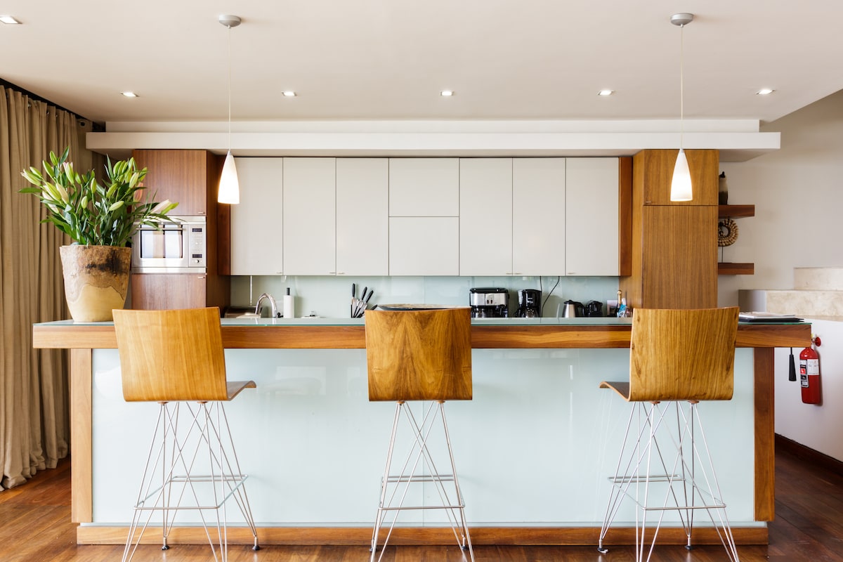 A modern kitchen bar area features three wooden stools positioned at a sleek counter. Under-cabinet lighting illuminates the minimalist cabinetry. A large potted plant adds a touch of greenery, while appliances and utensils are neatly arranged in the background.