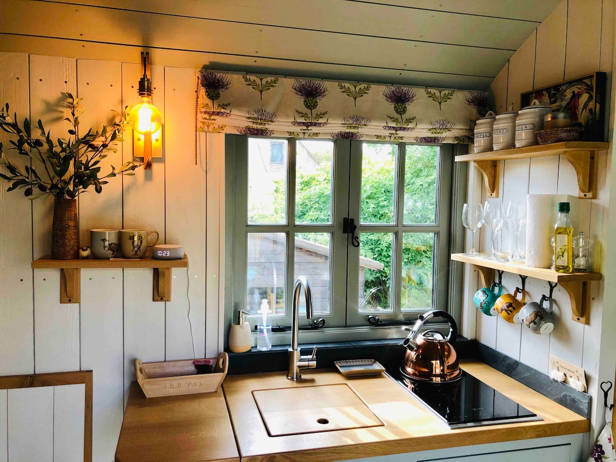 The kitchen area features a stylish layout with a wooden countertop and a Belfast sink. A kettle sits on the stovetop beside elegant glassware. Natural light enters through a window adorned with floral curtains, complemented by decorative shelves displaying cups and small plants.