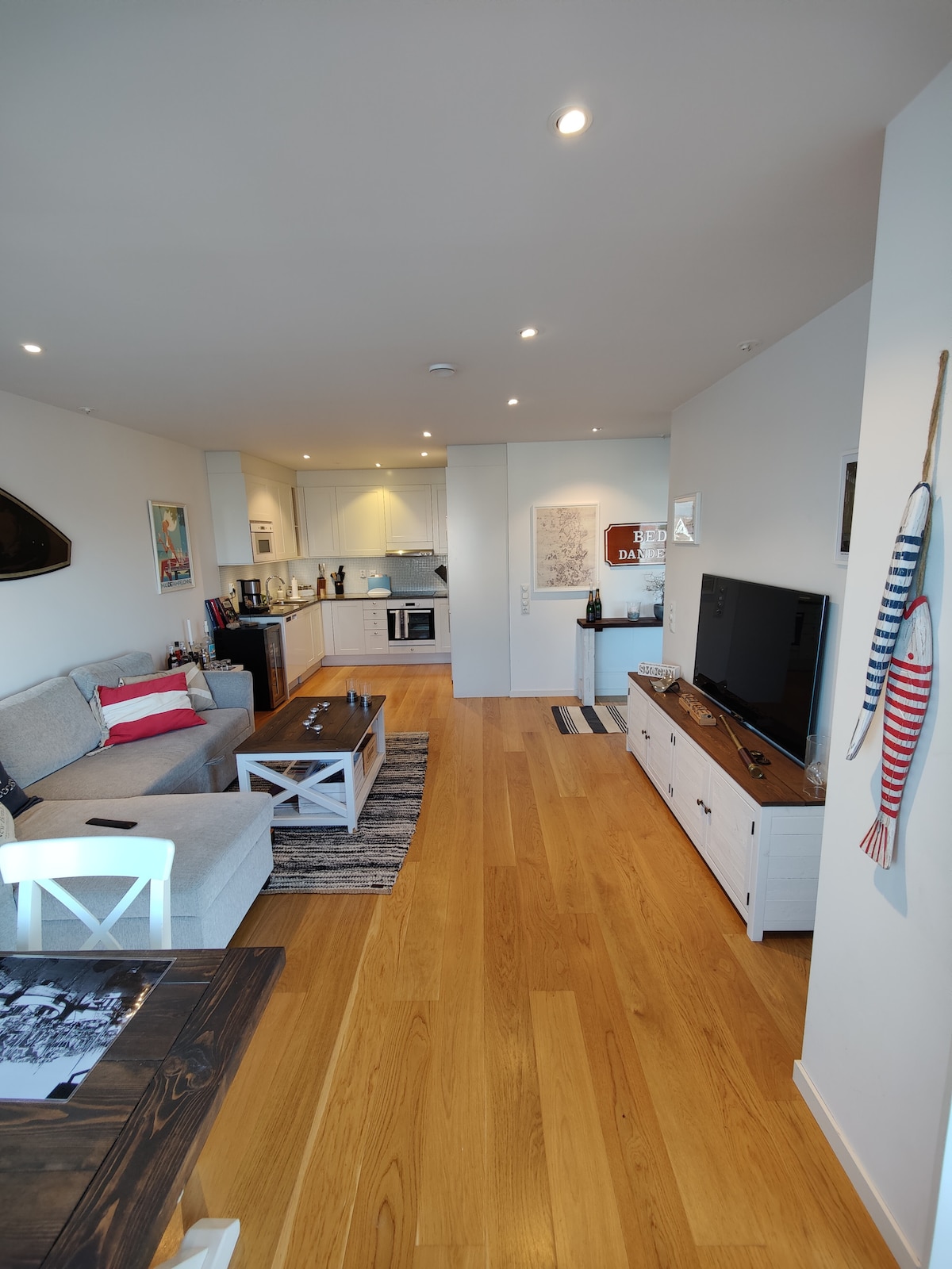 A modern living area is visible, featuring a comfortable sofa with accent pillows and a coffee table. The kitchen area, seen in the background, is equipped with white cabinetry and modern appliances. Natural light fills the space, highlighting the warm wooden flooring.