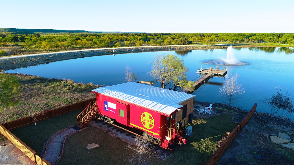 An aerial view captures the vibrant red train caboose nestled beside a serene lake, with a wooden dock extending towards a fountain in the water. Surrounding greenery reveals a spacious outdoor area with a winding path and a fenced private yard.