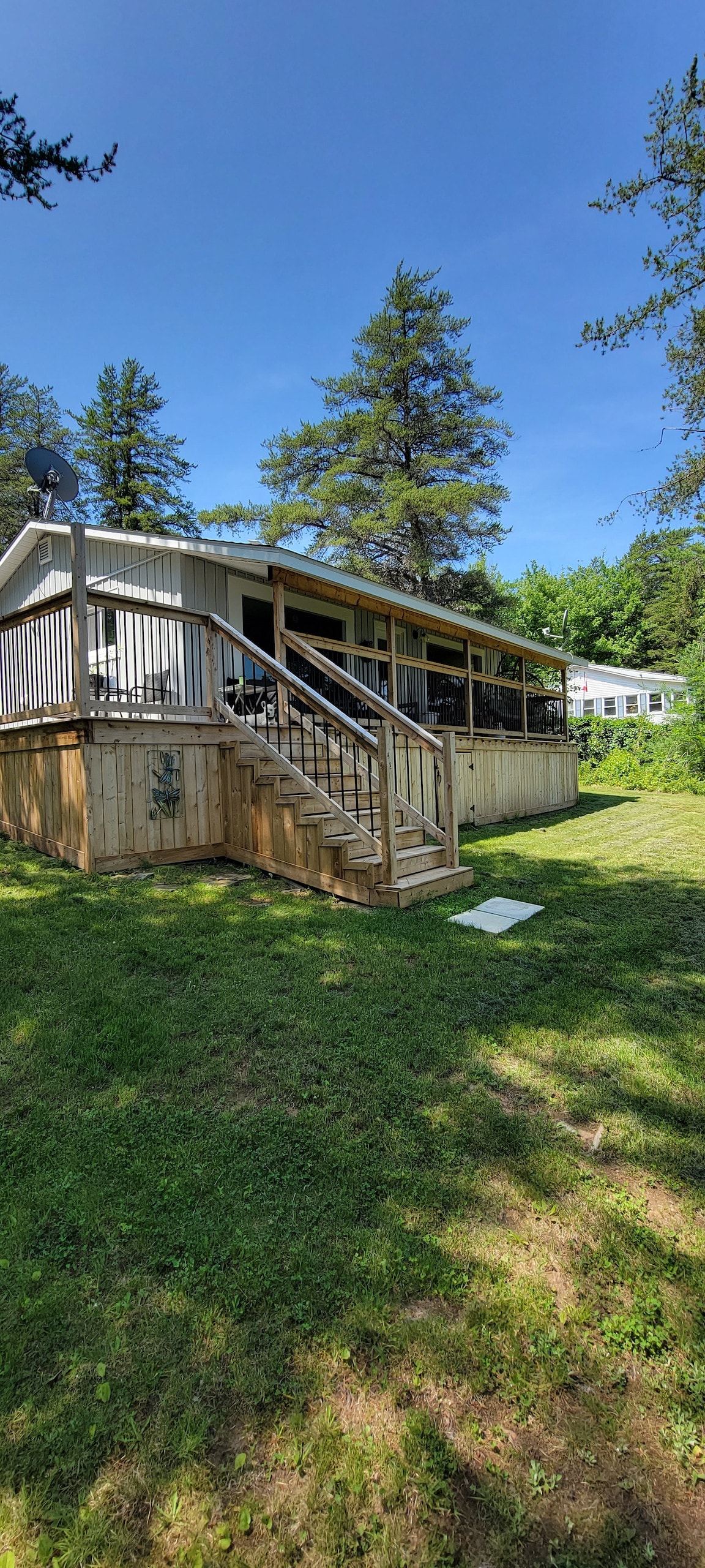 A cottage is set amid lush greenery, featuring a wooden deck with steps leading to the entrance. Dense trees provide shade while the lawn is kept tidy, offering a serene setting for relaxation. A satellite dish is visible, enhancing connectivity.