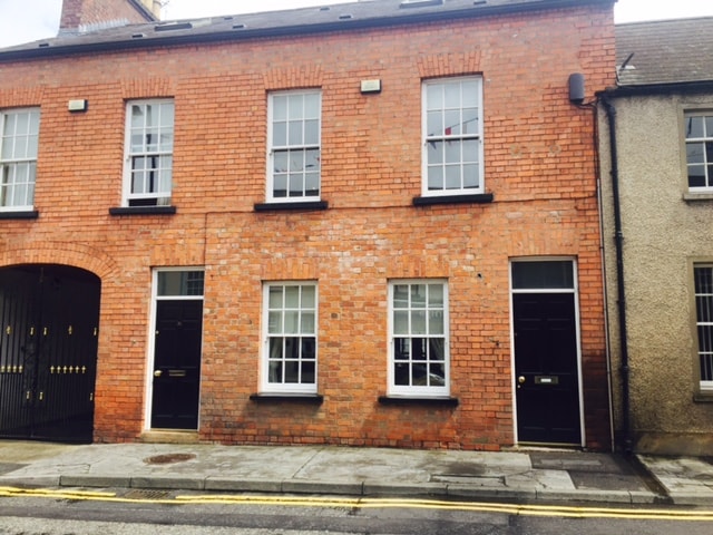 The exterior of a historic townhouse is presented, showcasing a red brick façade with white window frames. The entrance features a black door with a handle. Large windows allow natural light, and a paved pathway runs alongside the building.