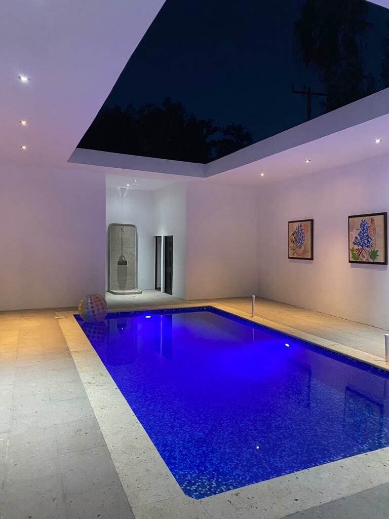 An indoor pool area is illuminated by soft lights, with water exhibiting a calming blue hue. Two wall-mounted artworks featuring blue flowers are positioned above the pool, enhancing the modern design. The ceiling features a skylight, allowing for starlit views at night.