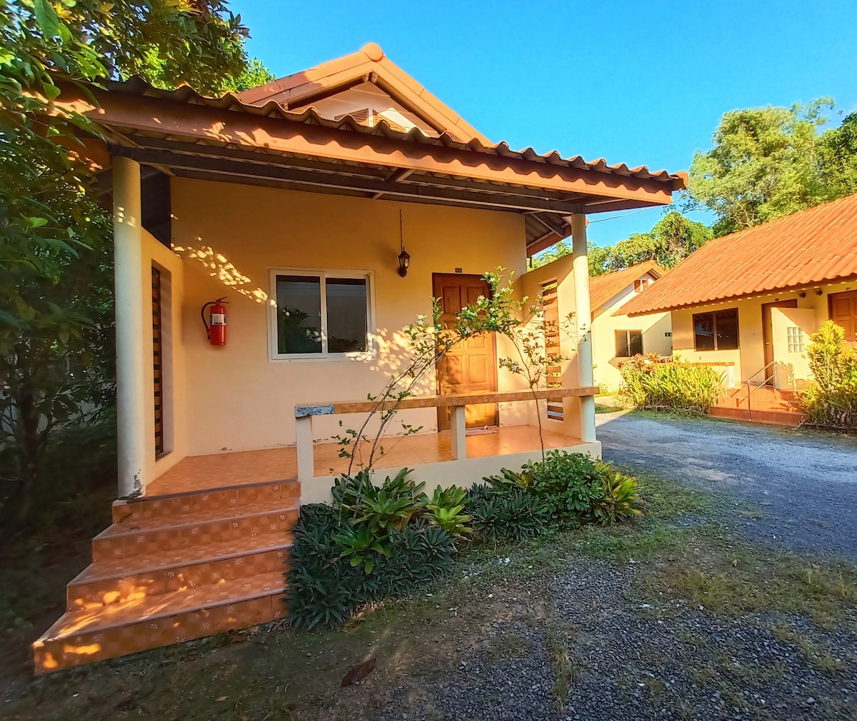 A single-story cabin with a welcoming entrance features a small porch and a front door framed by large windows. Lush greenery surrounds the property, and a gravel pathway leads to the cabin, enhancing its tranquil and secluded ambiance.